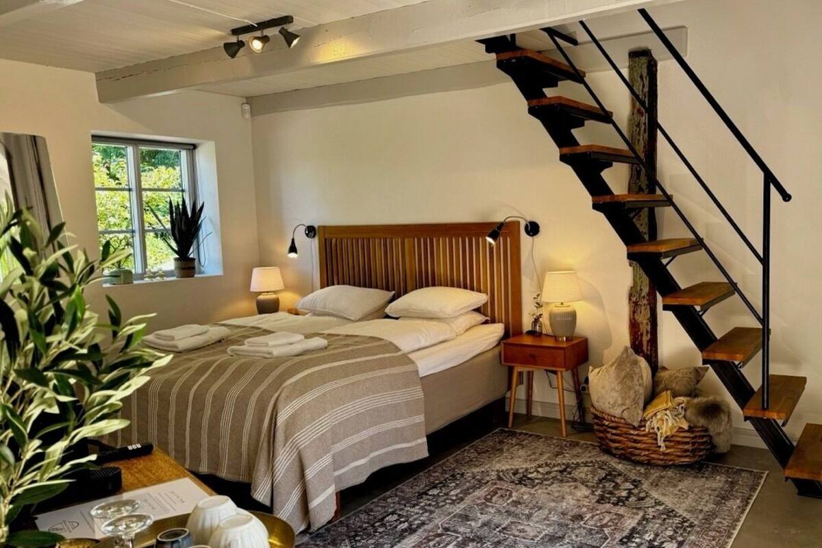 Ein Schlafzimmer mit Bett, Nachttischen, einer Lampe, einem Teppich, einer Treppe und einem Fenster mit Pflanzen.