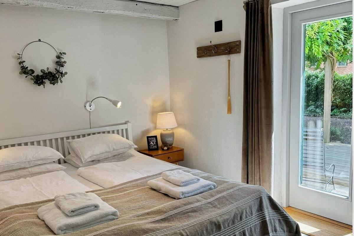 Ein Schlafzimmer mit einem Bett, Kissen, einem Nachttisch mit Lampe, einer Wandleuchte und Blick ins Freie durch ein Fenster.