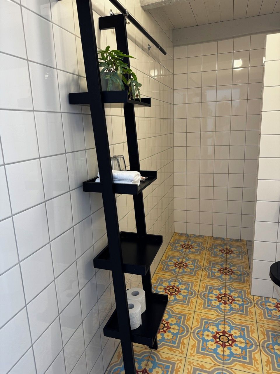 Ett badrum med vita kakelväggar, en svart metallhylla med en krukväxt, ett mönstrat golv och en toalett.