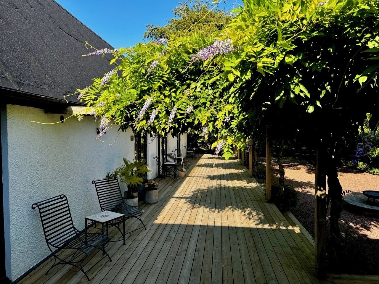 Een houten terras met buitenmeubilair en een pergola begroeid met bloeiende wijnranken.