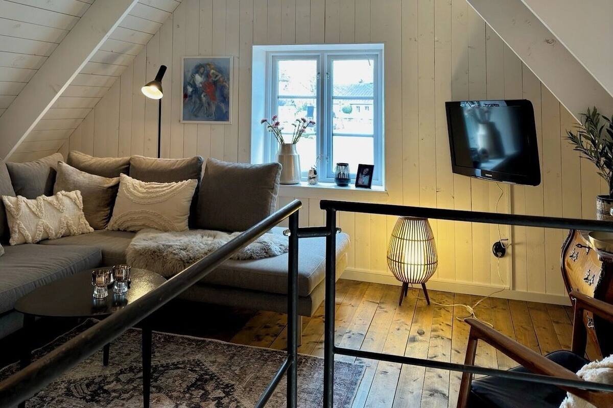 Ein gemütliches Wohnzimmer im Loft-Stil mit schrägem Dach, einer Couch mit Kissen, einem Couchtisch, einem an der Wand befestigten Fernseher und einem Fenster mit Ausblick.