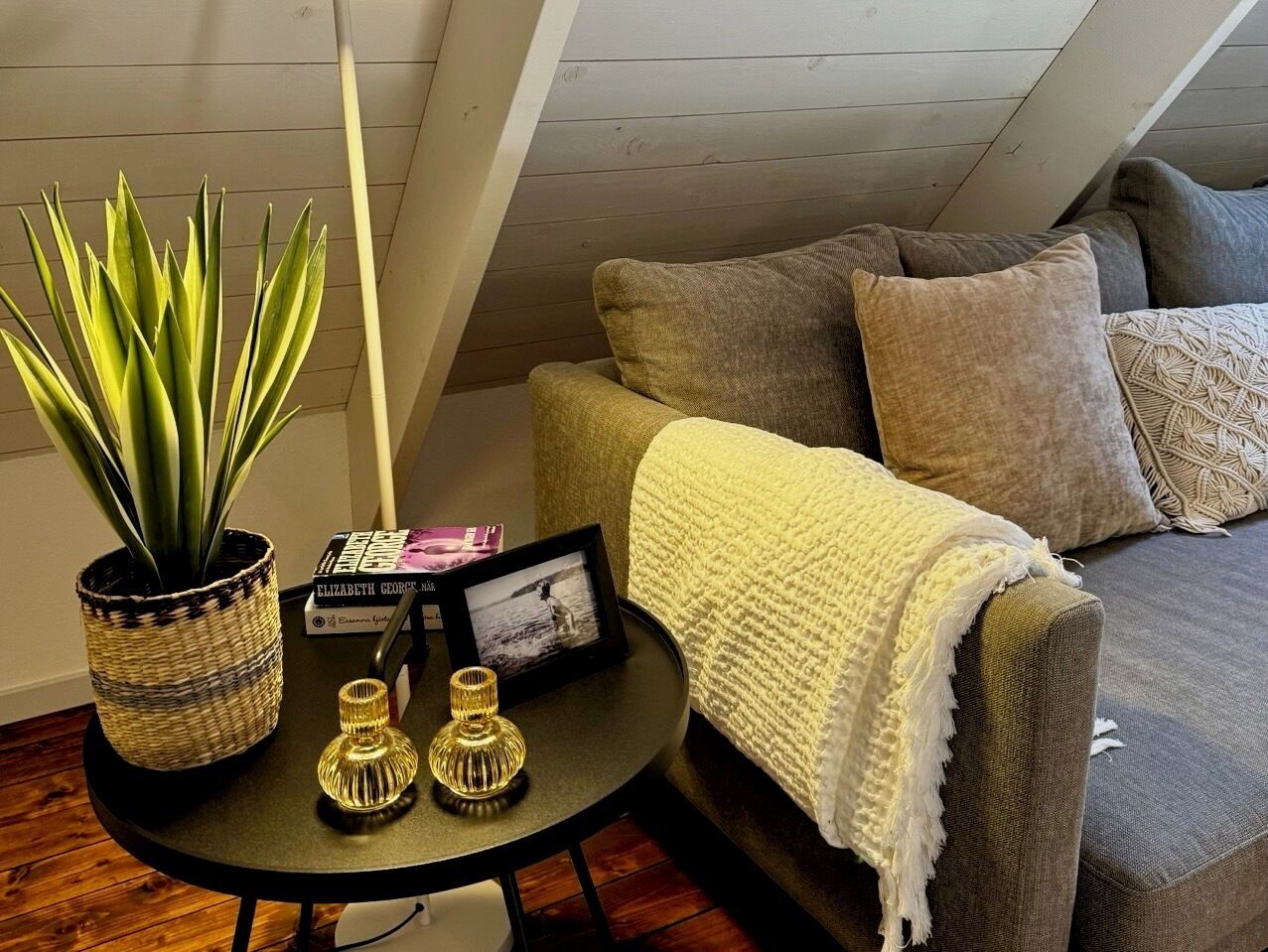 A cozy room with a grey sofa, a white throw blanket, and a small round table with a potted plant, a framed photo, and decorative vases.