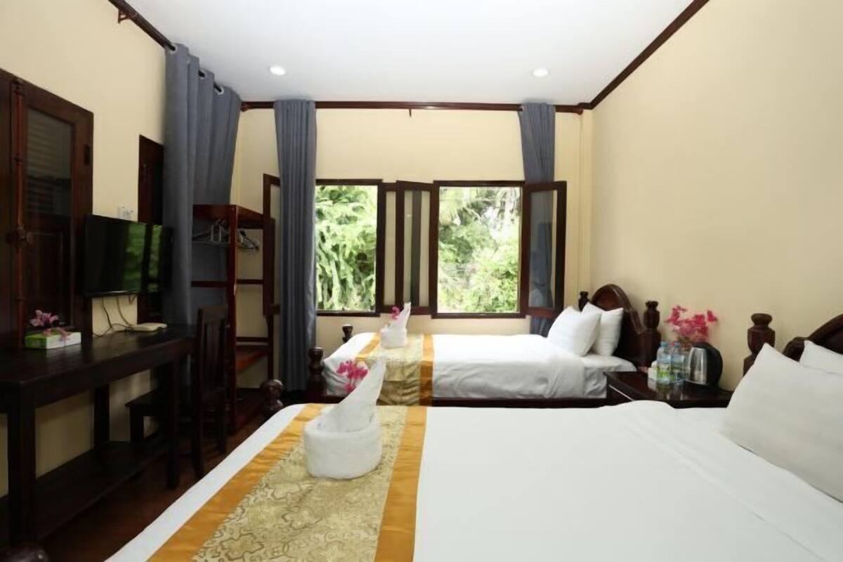A hotel room with two beds, a desk, a TV, and a window with a view of greenery.