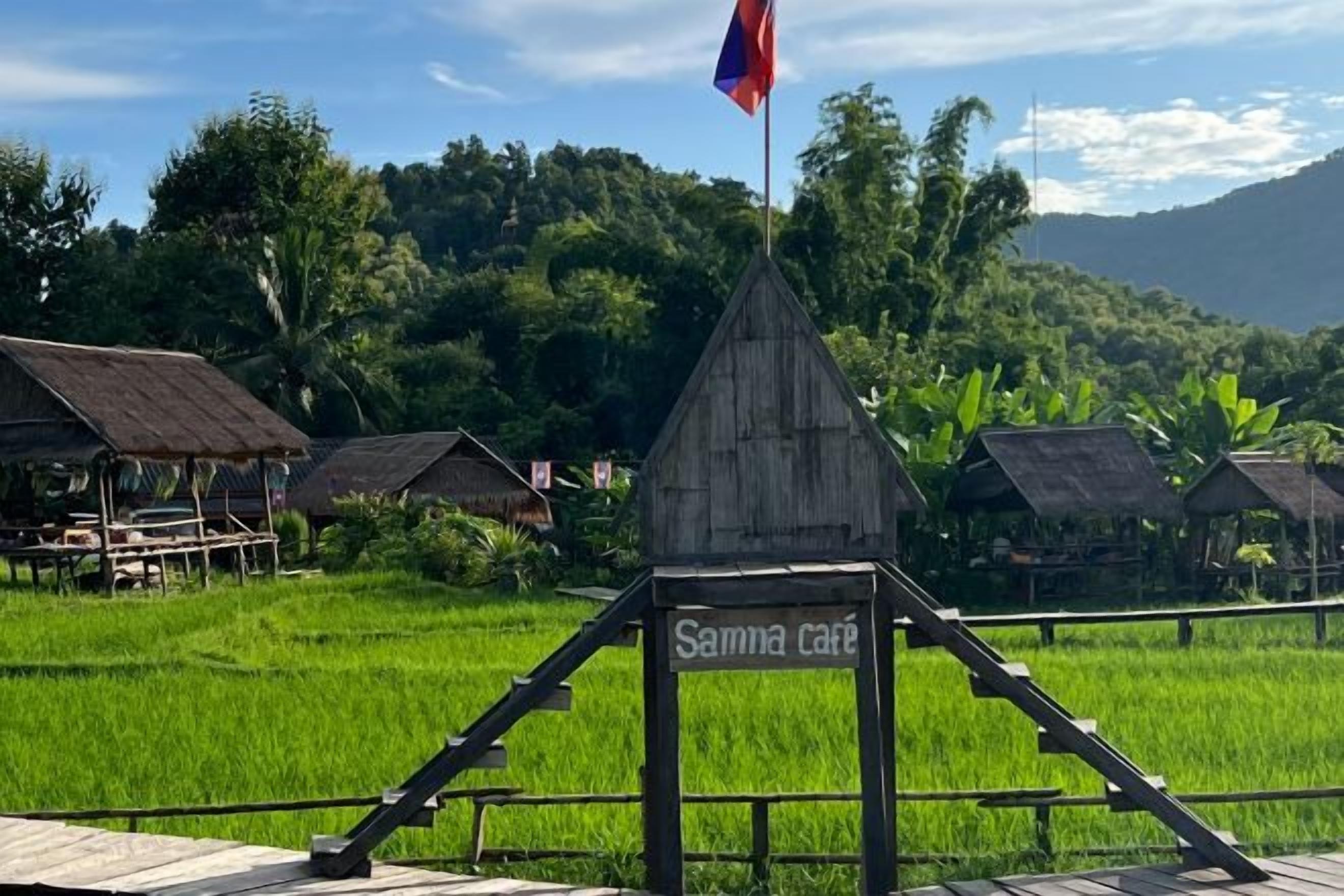 A rustic wooden structure with a sign reading 'Samma cafe' in a lush green landscape with rice fields and traditional huts.