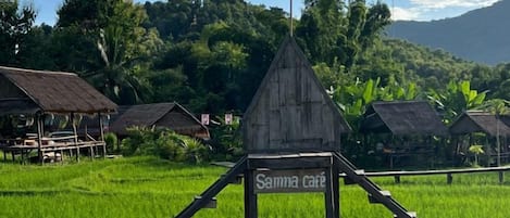 A rustic wooden structure with a sign reading 'Samma cafe' in a lush green landscape with rice fields and traditional huts.