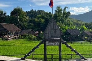 A rustic wooden structure with a sign reading 'Samma cafe' in a lush green landscape with rice fields and traditional huts. - We House Guest Hotel (Luang Prabang)
