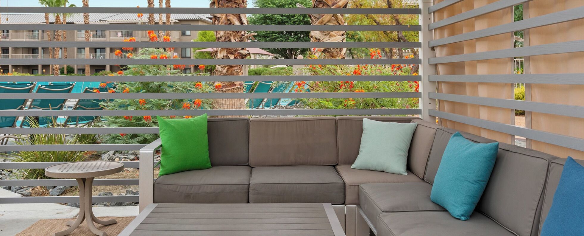 Un balcon avec un canapé gris, une petite table ronde et une vue sur un espace piscine agrémenté de chaises et de plantes.