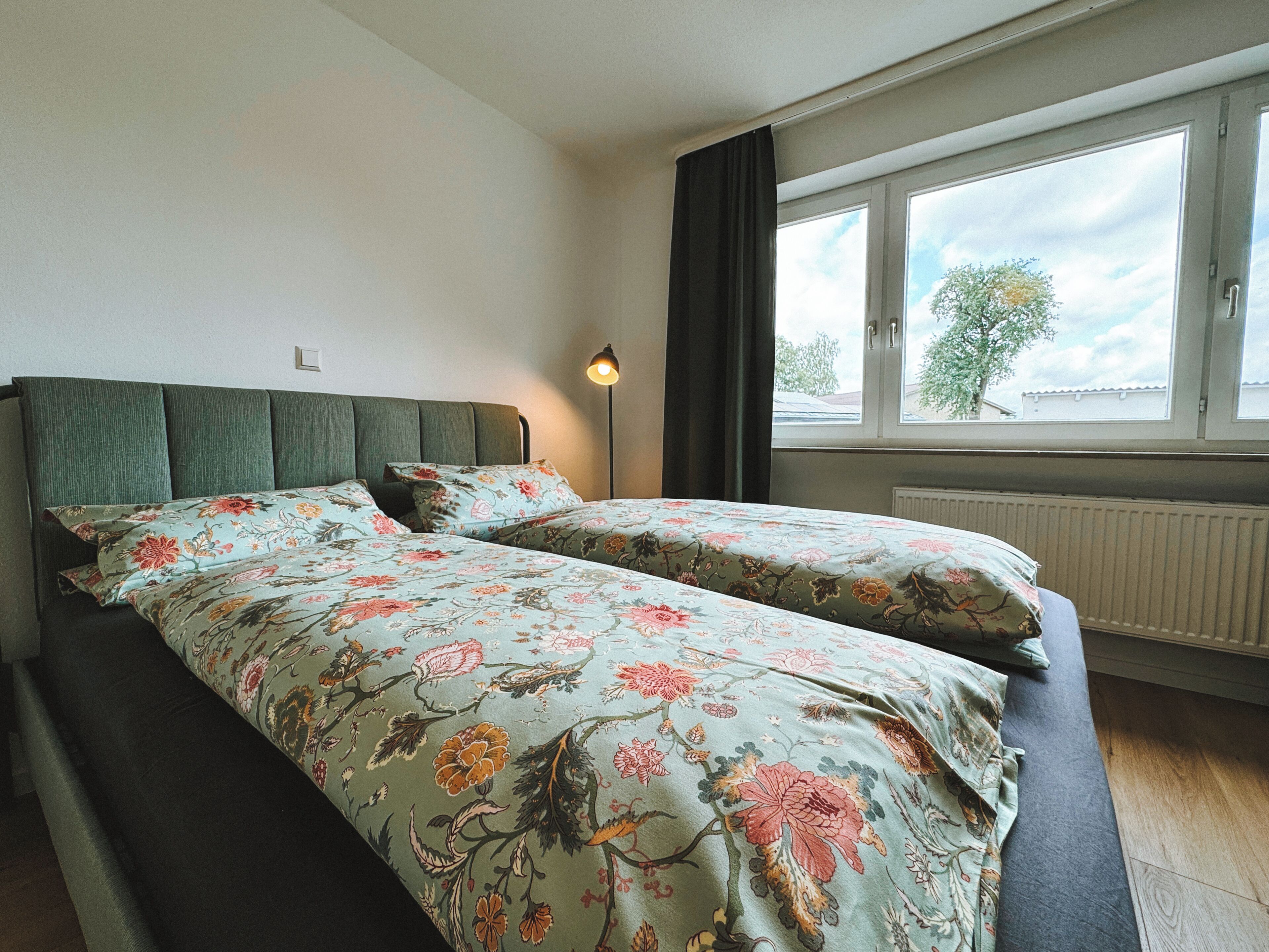 A double bed with floral bedding, a bedside lamp, and a window with a view of trees and buildings.