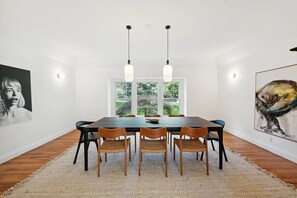 Un comedor con una mesa rectangular, seis sillas, dos lámparas colgantes, un ventanal con vistas a la vegetación y obras de arte en las paredes.