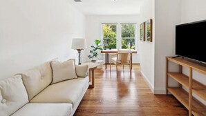 A modern living room with a sofa, a dining table with chairs, a TV on a wooden stand, and a large window with a view of trees. - Head of Pond House (Water Mill)