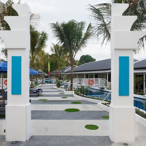 L’entrée d’un complexe hôtelier agrémentée de palmiers, d’une piscine et d’un espace abrité avec des parasols.