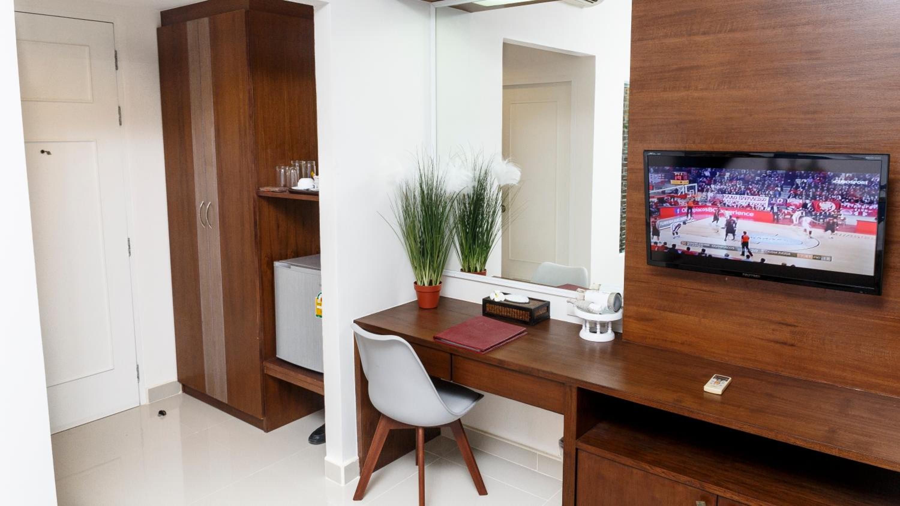 A hotel room with a wooden desk, a television mounted on the wall, a potted plant, and a white chair.