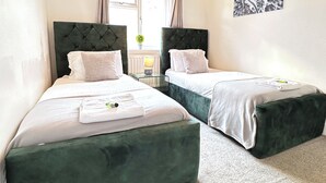 Une chambre d’hôtel avec deux lits, chacun doté d’une tête de lit recouverte d’un tissu vert, de draps blancs et d’une petite table avec une lampe placée entre les deux.
