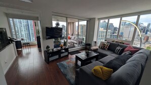 A modern living room with a large sofa, a coffee table, and a TV. There are large windows offering a city view. - Modern DT 2-Bed Condo, Large Balcony, Great View, Free Parking, A/C, EV charger (Vancouver)