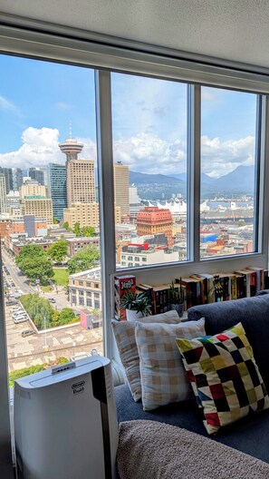 Ein Zimmer mit großem Fenster und Blick auf die Stadtlandschaft, einer Couch mit Kissen sowie einem Bücherregal mit Büchern.