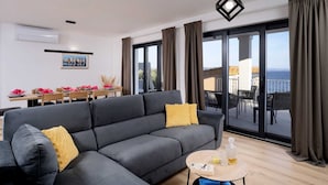 Un salon moderne avec un canapé gris, une petite table ronde sur laquelle se trouvent une bouteille et des verres, et un coin repas avec une table dressée pour quatre personnes.