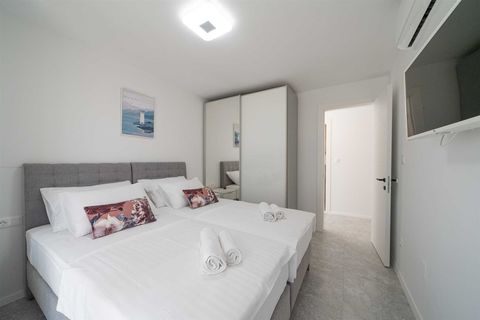 Een tweepersoonsbed met witte lakens, twee kussens en opgerolde handdoeken. Er is een grijs hoofdbord, een nachtkastje met een lamp en een aan de muur bevestigd kunstwerk.