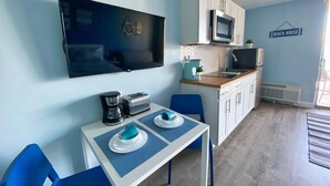 A compact kitchen with a microwave, toaster, and coffee maker. There is a dining table with blue chairs and a TV mounted on the wall. - NEW Amazing Ocean Front King Bed Studio in Daytona! (Daytona Beach)