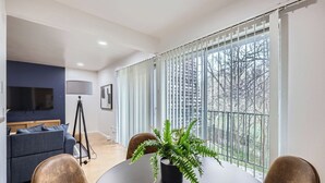 A modern living room with a dark blue sofa, a round dining table with chairs, a potted fern, and a large window with vertical blinds.