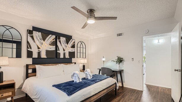 A modern hotel room with a large bed, a ceiling fan, a desk with a chair, and a large abstract painting on the wall.
