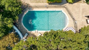 An aerial view of a rectangular swimming pool with a clear blue water surface, surrounded by a paved deck and bordered by trees.