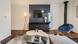 A modern living room with a white bench, a blue chair, a black entertainment unit, and a black fireplace.