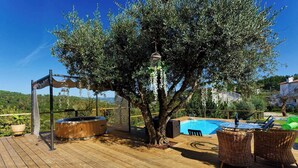 Een houten terras met een jacuzzi, een zwembad, rieten meubels en een pergola met hangende lampen.