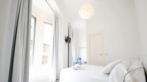 A bright, white hotel room with a large bed, a bedside table, a TV, and a view of the outside. - Dingdong Express Hostel (Barcelona)