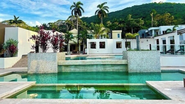 A modern resort with a central pool, surrounded by white buildings and palm trees, set against a backdrop of green hills. - Azalia Palms: New Oceanview Villa, 15 Min from MBJ Airport (St. James Parish)