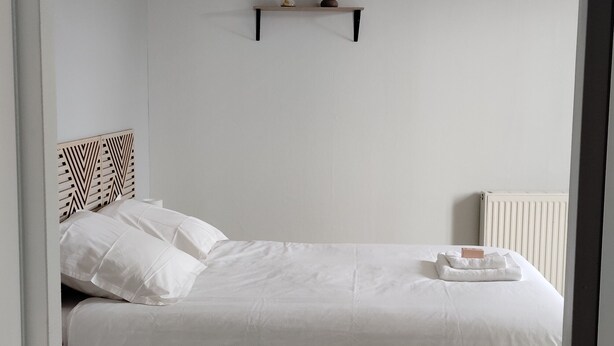 A minimalist bedroom with a bed, a small shelf, a radiator, and a woven light fixture. - Studio+outside Ribérac town center2 (Ribérac)