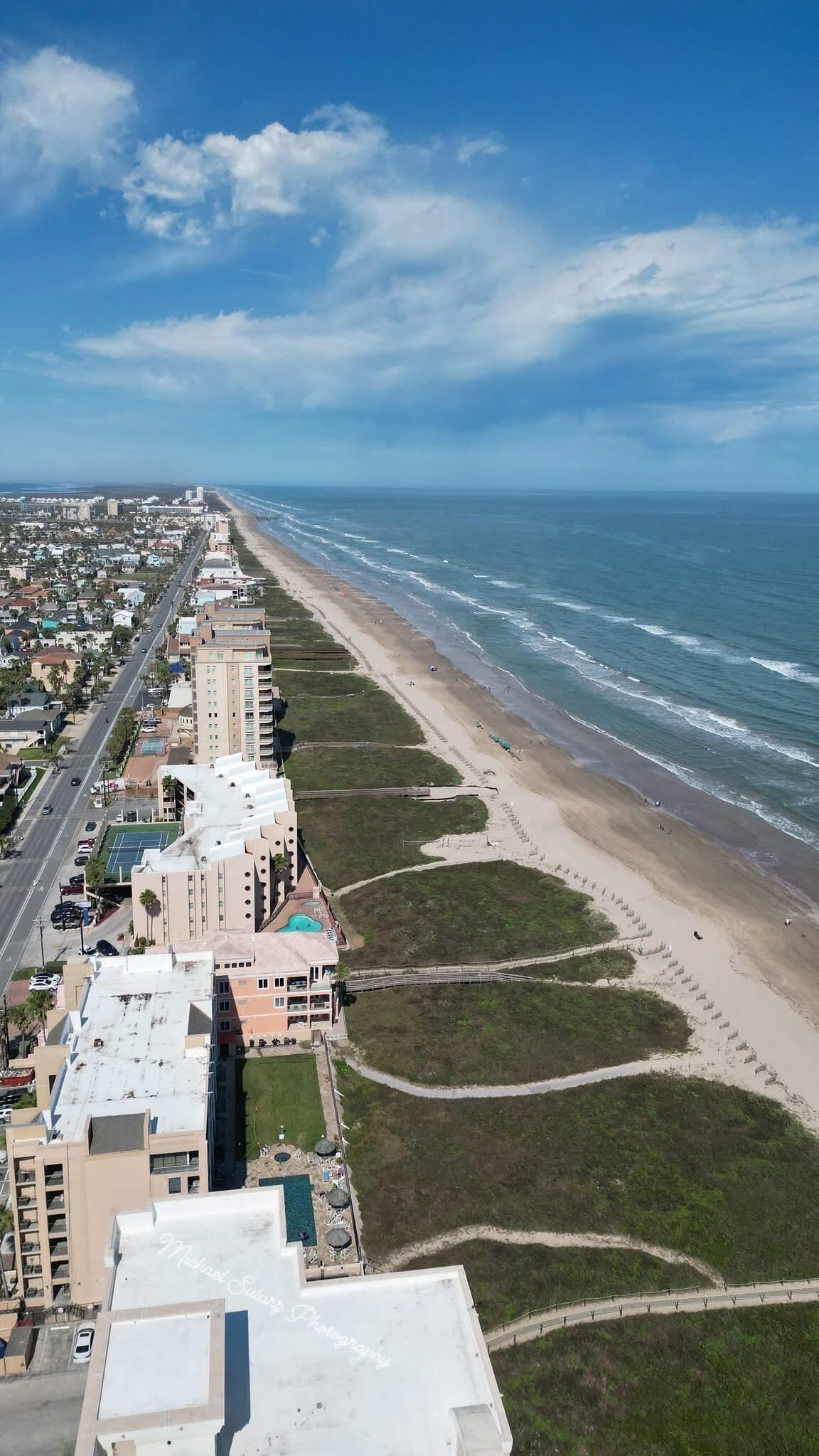 An aerial view of a coastal area with a beach, buildings, and a road.