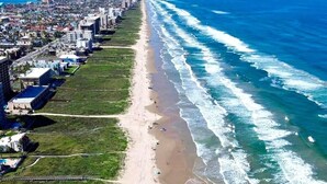 Vista aérea de una ciudad costera con una playa extensa, una zona urbana densa con edificios y un cielo azul despejado.