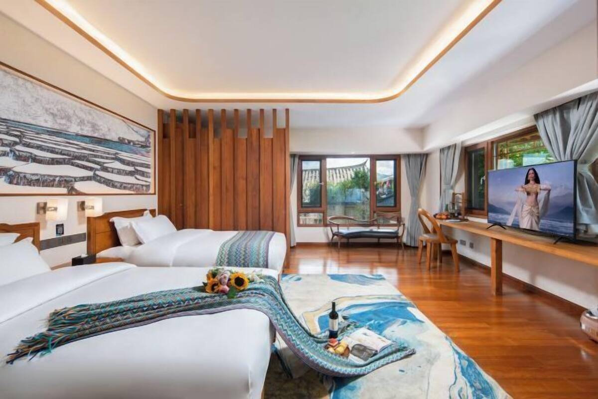 A modern hotel room with a large bed, a desk with a TV, and a view of the outdoors through large windows.