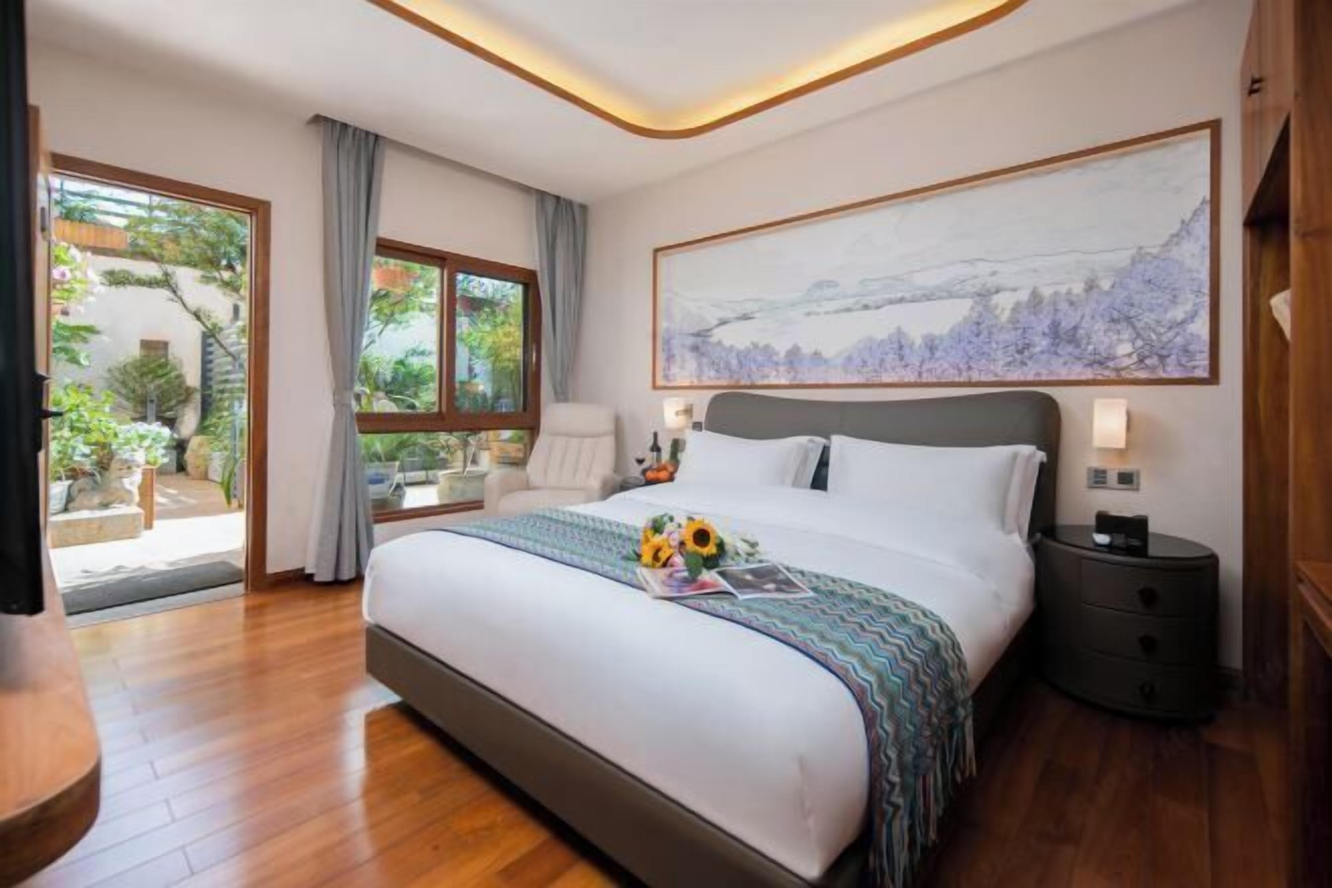 Une chambre avec un grand lit, une table de chevet, une chaise, une fenêtre donnant sur un paysage verdoyant et un tableau accroché au mur.
