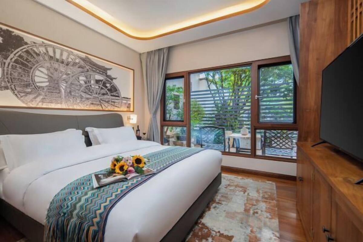 A modern hotel room with a large bed, a television, a window with a view of greenery, and a decorative wall piece.