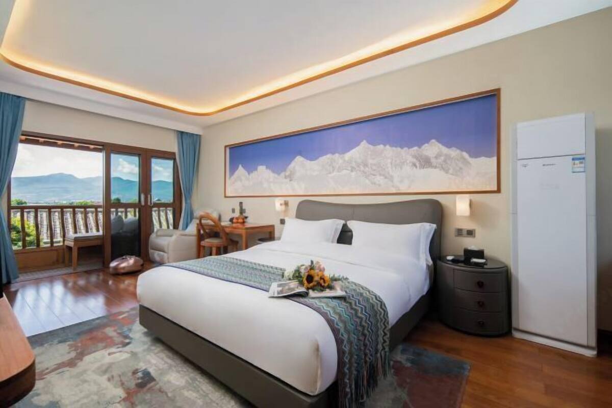 A hotel room with a large bed, a desk, a chair, a nightstand, and a view of mountains through the window.