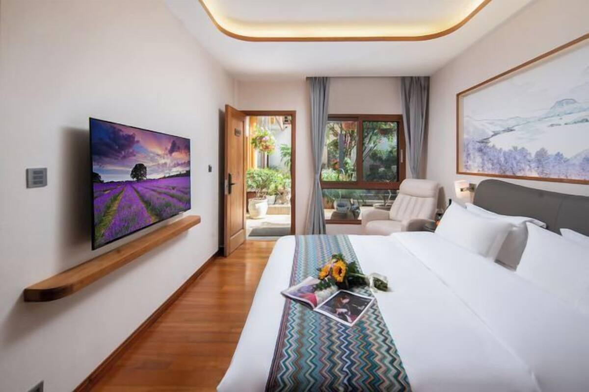 A hotel room with a bed, a TV mounted on the wall, a window with a view of greenery, and a painting on the wall.