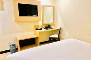A hotel room with a bed, a desk with a chair, a television, a mirror, and a trash bin. - Jakarta City Hotel (West Jakarta)