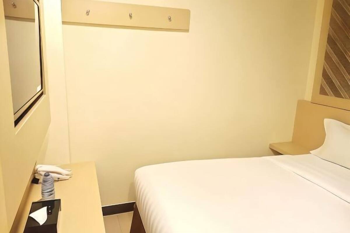 Une chambre d’hôtel avec un lit, une table de chevet, un miroir et une étagère fixée au mur.