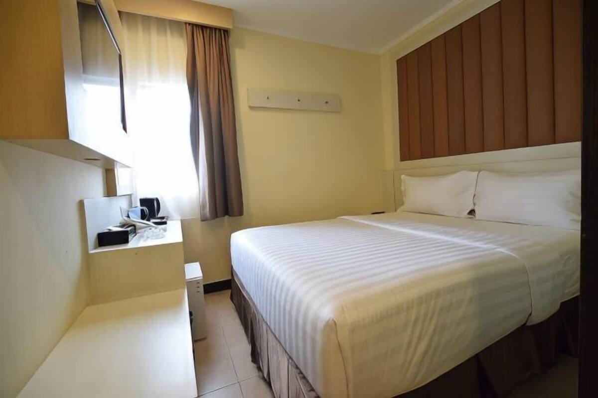 Une chambre d’hôtel comprenant un lit, un bureau avec une chaise, une fenêtre avec des rideaux et un luminaire fixé au mur.