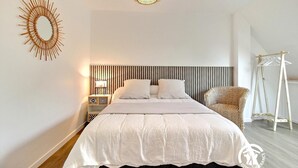 A modern bedroom with a large bed, a bedside table with a lamp, a decorative wall piece, and a hanging light fixture. - Marine calm - QUIBERON (QUIBERON)