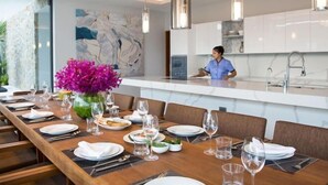 A modern dining area with a long wooden table set for a meal, a kitchen in the background, and a large abstract painting on the wall. - Exclusive Sea Renity A Beachfront Villa Retreat Sleeps 12 Adults and 6 Kids (Koh Samui)