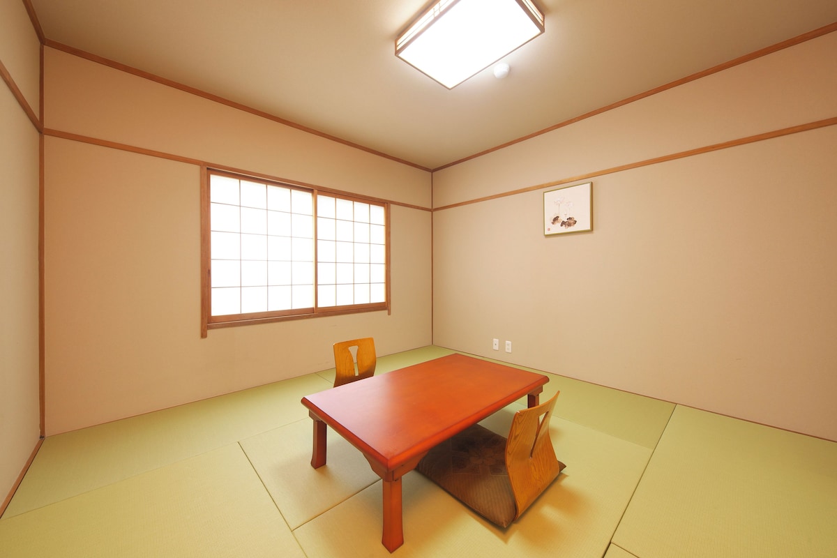 A room with tatami flooring, a wooden table, two chairs, a window with a grid pattern, and a framed picture on the wall.