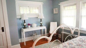 A bedroom with a bed, a white console table with lamps, a radiator, and a window with curtains. - The Trillium House at DePauw - 4/5 BR 3.5 BA (Greencastle)