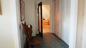 A hallway with a wooden chair, a door leading to a room with a bed and a nightstand, and a closet with white shutters. - The Trillium House at DePauw - 4/5 BR 3.5 BA (Greencastle)