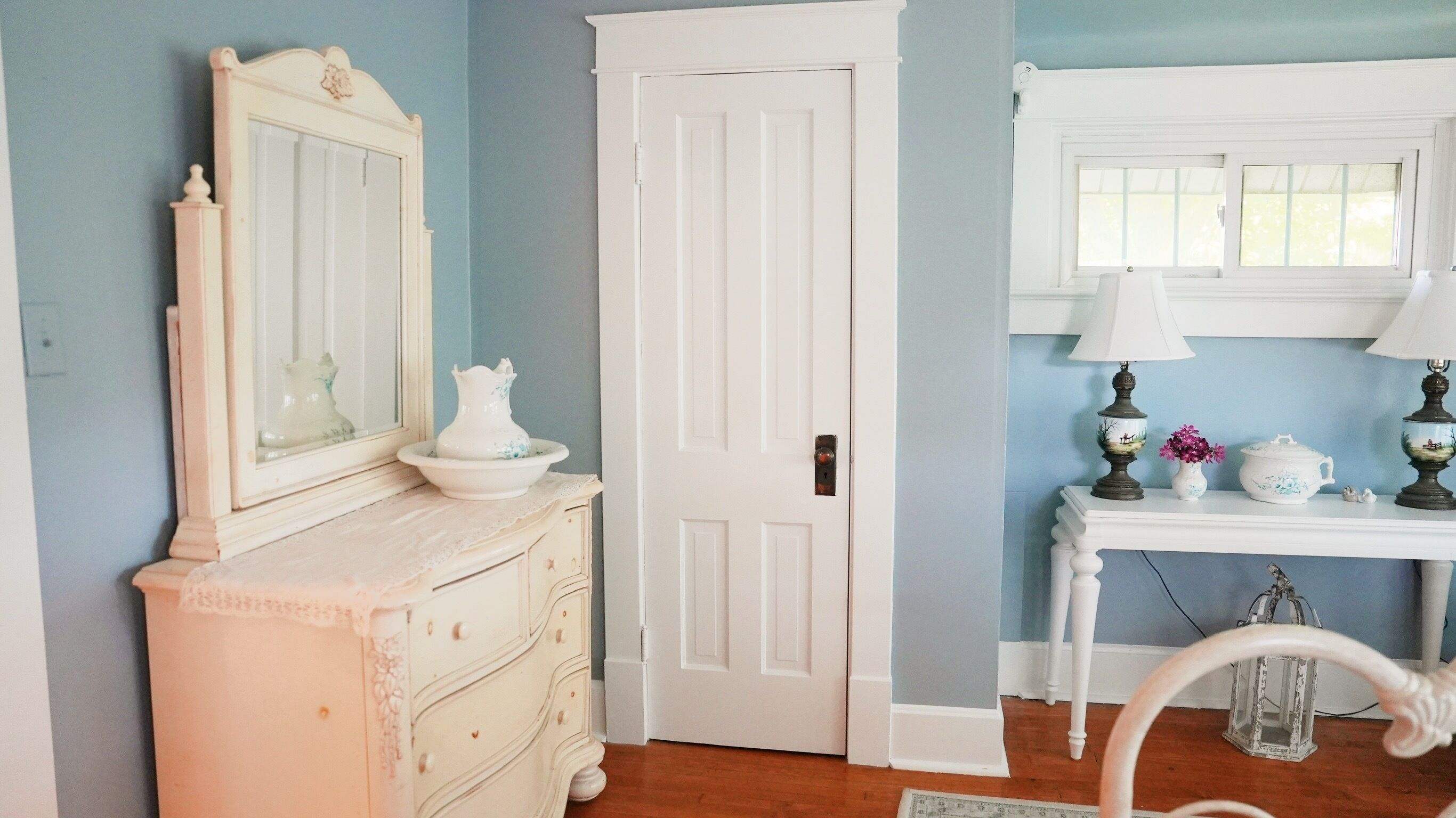 Une pièce avec une commode blanche, une porte, une console ornée d’objets décoratifs et une fenêtre à cadre blanc.