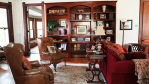 A traditional living room with leather armchairs, a red sofa, and a wooden bookshelf filled with books and decorative items. - The Trillium House at DePauw - 4/5 BR 3.5 BA (Greencastle)
