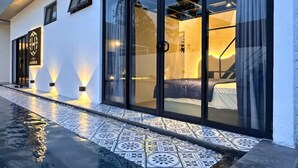 Exterior moderno de un hotel con entrada de vidrio, piso de baldosas y una habitación visible con cama y cortinas.