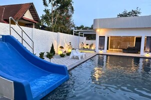 Una piscina con un tobogán azul, sillas reclinables blancas y un edificio moderno con grandes ventanales.