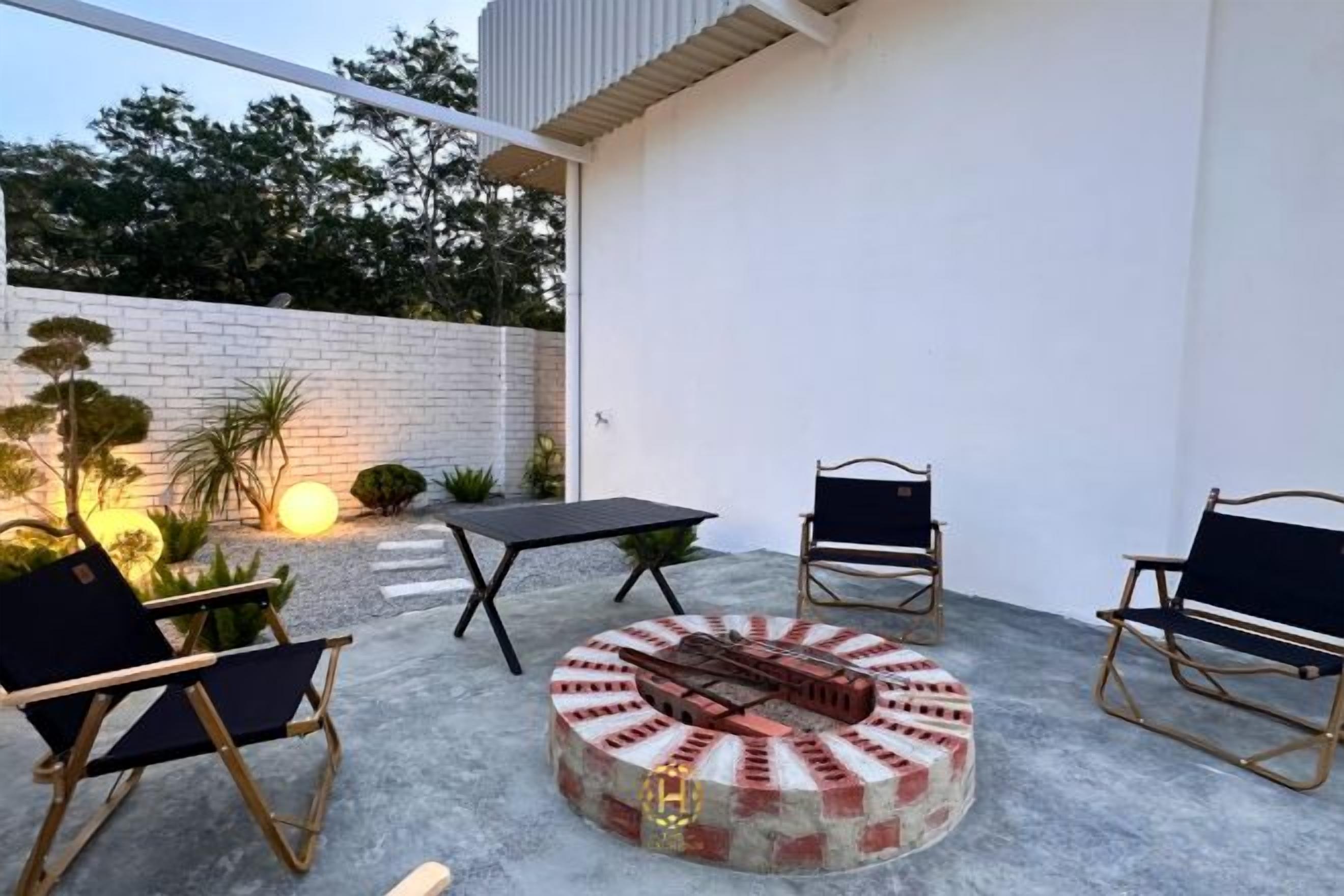 An outdoor patio with a round fire pit, two chairs, a table, and a brick wall.